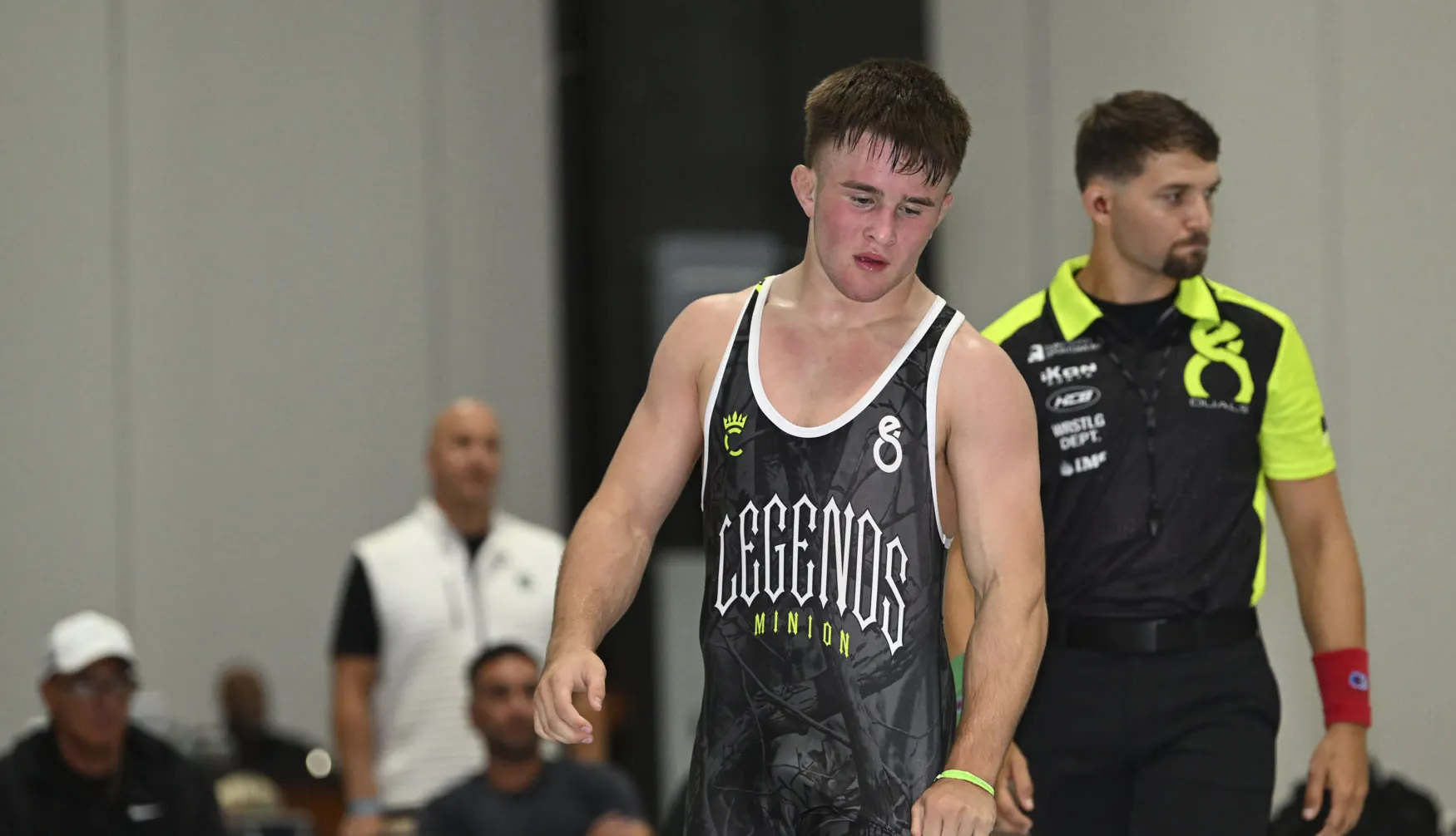 Wrestler in a black Legends singlet walking off the mat beside the referee