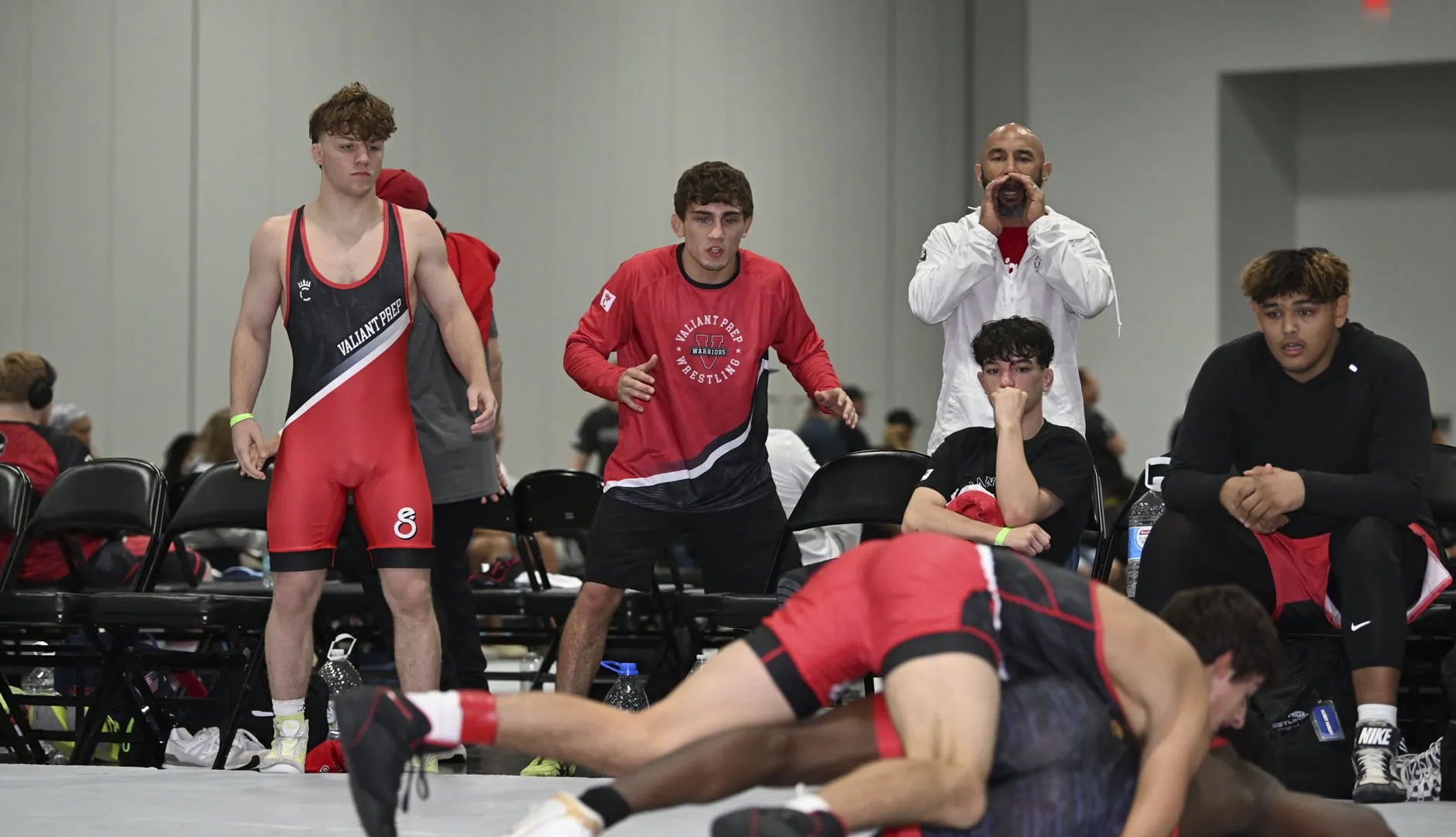 A wrestler in a red and black singlet driving for a pin as his team watches
